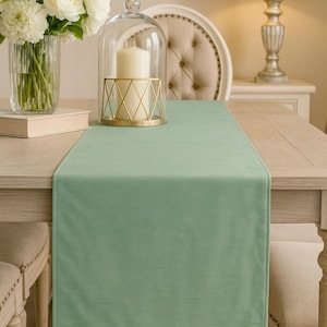 May include: A sage green table runner is draped across a light wood table. A glass cloche with a candle sits on the runner, next to a vase of white flowers. A lamp and chair are in the background.