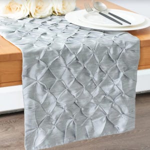 May include: A silver table runner with a quilted pattern. The runner is made of a soft, silky fabric and has a subtle sheen.