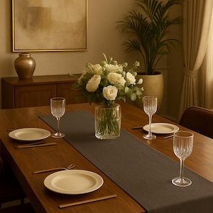 May include: A dining table set for a meal, featuring a dark gray table runner, white plates, and clear wine glasses. A vase of white roses sits in the center, with a chandelier overhead. The table is made of wood.