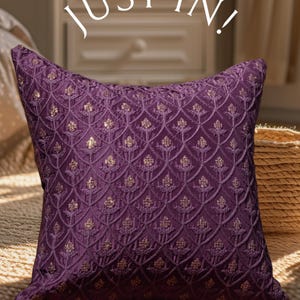 Peut inclure: Un coussin de couleur pourpre avec un motif floral brodé en or. Le coussin est posé sur une surface en tissu brun. Le texte "Just In!" est écrit en blanc au-dessus du coussin.