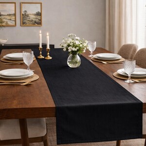 May include: A long, rectangular black table runner made of fabric, centered on a wooden dining table. The runner features a smooth, solid-colored surface with clean, straight edges, providing a dark contrast against the natural wood grain.