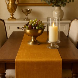 May include: A dining table set with a mustard-colored table runner, a brass bowl filled with grapes, and a lit candle in a glass holder. A gold-framed painting and decorative items are in the background.