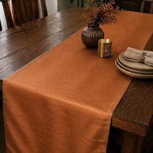 May include: A solid burnt orange rectangular table runner made of a textured fabric. The runner is draped lengthwise across a dark brown wooden table, providing a smooth surface for decorative items like a vase, a candle holder, and plates.