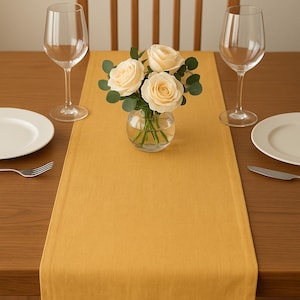 May include: A dining table setting with a mustard yellow table runner. Two wine glasses, white plates, and silverware are set on the wooden table. A small glass vase holds a bouquet of cream-colored roses.