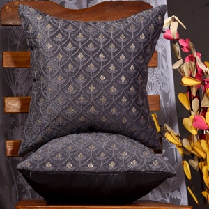 May include: Two gray throw pillows with a gold embroidered pattern. The pillows are stacked on top of each other and are sitting on a wooden chair.
