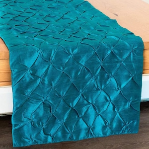 May include: Teal-colored table runner with a textured, quilted pattern. The runner is made of a shiny fabric and is draped over a wooden surface. The quilted design creates a diamond-like pattern across the fabric.