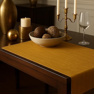 May include: A dining table set with decorative items. A gold-colored vase, candle holders, and a bowl with decorative spheres are arranged on a mustard-colored table runner. A wine glass and a candelabra with lit candles are also present.