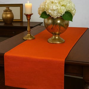 May include: An orange table runner is centered on a dark brown wooden table. A gold-colored candle holder with a lit candle and a gold vase with white flowers are on the table. A gold decorative jar and framed mirror are in the background.