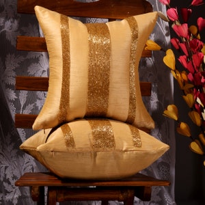 May include: Two square, gold-colored throw pillows with vertical beaded stripes. The pillows are stacked on a wooden chair. The background includes a floral curtain and decorative flowers in shades of red, yellow, and pink.