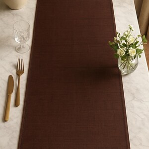 May include: A dark brown table runner is centered on a marble table. A glass of water, a fork, and a knife are on the left. A small vase of white flowers is on the right. The runner is made of a textured fabric.