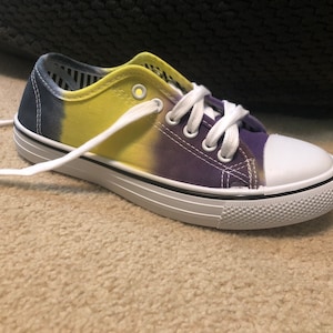 May include: A pair of white canvas low-top trainers with a tie-dye pattern in shades of blue, yellow, and purple. The laces are white.