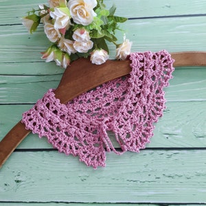 Puede incluir: Un collar de crochet rosa con un delicado patrón de encaje, mostrado en una percha de madera sobre un fondo verde claro.