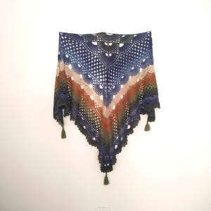 May include: A crocheted shawl with a triangular shape and a multi-colored gradient pattern. The shawl is made with blue, green, brown, and beige yarn. It has a fringe border and tassels at the corners.