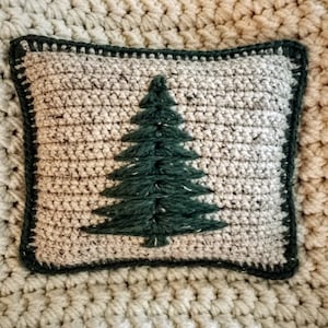 May include: A crocheted pillow with a green embroidered pine tree design. The pillow has a beige background and a dark green border.