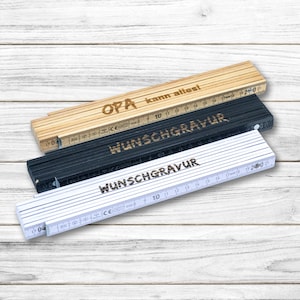 Könnte beinhalten: Drei Holz-Klapplineale mit dem Text "OPA kann alles!" auf dem oberen Lineal und "WUNSCHGRAVUR" auf den beiden unteren Linealen. Die Lineale liegen auf einer hellen Holzoberfläche.