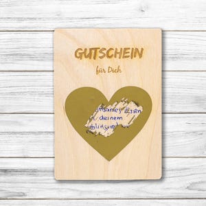 Könnte beinhalten: Geschenkgutschein aus Holz mit den deutschen Worten "Gutschein für Dich" oben eingraviert. In der Mitte befindet sich eine goldene Herzform mit handschriftlichem Text. Der Hintergrund ist eine helle Holzmaserung.