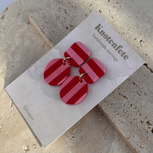 Könnte beinhalten: Ein Paar handgefertigte Ohrringe mit pinkfarbenen und roten Streifenmustern. Die Ohrringe werden auf einer weißen Karte mit dem Text "Knotenfête handmade earrings" und "Schwerin" präsentiert.