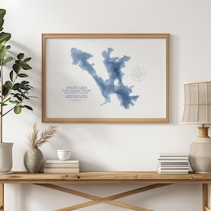 May include: A framed print of Aylen Lake, South Algoma Township, with a detailed blue topographical map. The print includes a compass rose and text with coordinates and measurements. The frame is a light brown color.