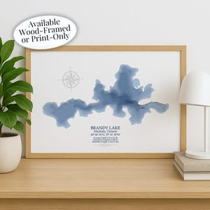 May include: A framed print of Brandy Lake in Muskoka, Ontario, with a detailed blue topographic map. The artwork includes a compass rose and text indicating the lake's location, average depth of 11 ft, and shoreline length of 7 miles. The frame is light wood.