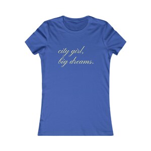 Peut inclure: Un t-shirt bleu roi à manches courtes avec le texte "city girl, big dreams." imprimé en police cursive jaune clair au centre de la poitrine. Le vêtement présente une encolure ronde et une coupe standard avec des manches courtes.