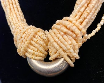 May include: A multi-strand necklace made of small, cream-colored shell beads. The necklace is knotted at the ends and features a silver clasp with the inscription "D.C. 1952".