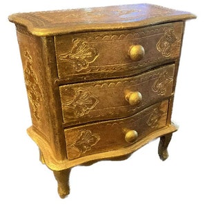 Puede incluir: Un pequeño cofre de cajones de madera pintado de oro con tres cajones. Cada cajón tiene un diseño floral decorativo y una perilla redonda.