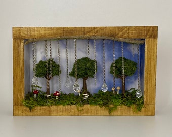 May include: A miniature forest scene in a wooden frame with a blue sky backdrop.  The scene features three small trees with green moss, red mushrooms, and silver chains with crystal drops hanging from the top of the frame.