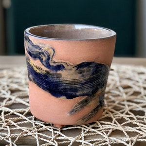 May include: A handmade ceramic cup. The exterior is peach-coloured with a dark blue and yellow painted design. The cup has a cylindrical shape with a slightly flared rim and sits on three small feet. The interior is a darker brown colour.