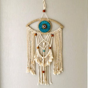 May include: A white macrame wall hanging with a blue evil eye charm and wooden beads. The hanging is in the shape of an eye and has a fringe at the bottom.