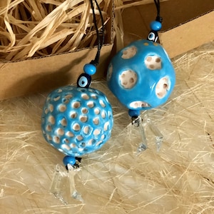 Puede incluir: Dos adornos de cerámica azul con detalles blancos y una cuerda negra que cuelga de ellos. Cada adorno tiene un amuleto de ojo turco azul en la cuerda. Los adornos cuelgan de una caja marrón con relleno de paja.