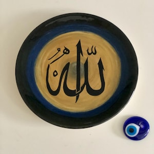 May include: A round ceramic plate with a blue and black border and a gold center. The word "Allah" is written in black calligraphy on the gold center. A small blue evil eye charm is in the lower right corner of the image.