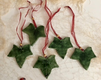 May include: Five green ceramic ivy leaf ornaments with red ribbon hangers. The leaves have a detailed texture and are hanging on a white background.