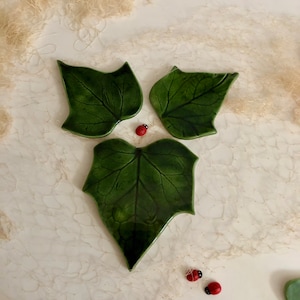 May include: Three green ceramic leaf-shaped plates with a realistic leaf design. The plates are arranged in a triangular shape with two plates at the top and one larger plate at the bottom. There are two small red ladybugs on the white surface below the plates.