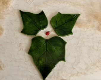 May include: Three green ceramic leaf-shaped plates with a realistic leaf design. The plates are arranged in a triangular shape with two plates at the top and one larger plate at the bottom. There are two small red ladybugs on the white surface below the plates.