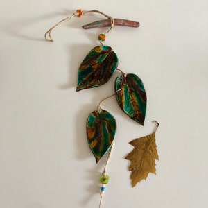 May include: A handcrafted wall hanging featuring three ceramic leaves with teal and brown hues, strung on twine. The leaves are accented with green and orange beads, and a small, dried leaf is also included.