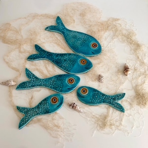 Pode incluir: Cinco ornamentos de cerâmica em forma de peixe turquesa, cada um apresentando um padrão floral detalhado em relevo no corpo e um olho circular proeminente com um centro escuro. Os peixes estão dispostos em grupo, exibindo o seu acabamento liso e brilhante.