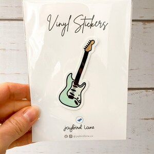 May include: Vinyl sticker of a light blue electric guitar with black and white details. The sticker is packaged in a clear plastic sleeve with the text "Vinyl Stickers" and "jaybird lane" printed on the white card.