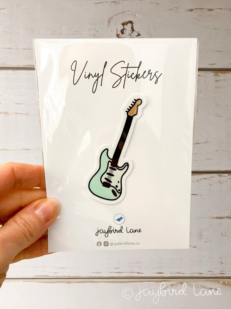 May include: Vinyl sticker of a light blue electric guitar with black and white details. The sticker is packaged in a clear plastic sleeve with the text "Vinyl Stickers" and "jaybird lane" printed on the white card.