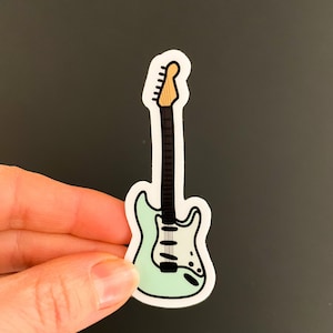 May include: A sticker of a light blue electric guitar with a white pickguard and a brown neck. The sticker is on a dark gray background.