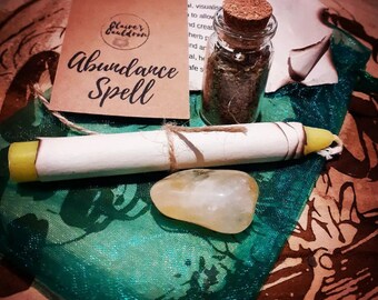 May include: An 'Abundance Spell' kit includes a small glass jar with a cork stopper, a rolled parchment, a yellow candle, a yellow crystal, and a small tag. The items are arranged on a green fabric with a floral pattern.