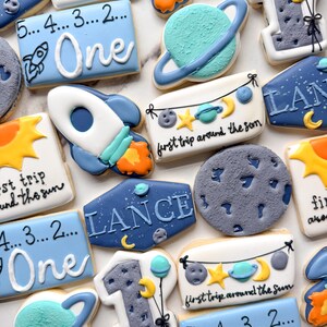 May include: A collection of space-themed sugar cookies featuring rockets, planets, moons, and the number one. Several cookies display the text "first trip around the sun" or "5... 4... 3... 2... One" with illustrations of stars, suns, and rockets.