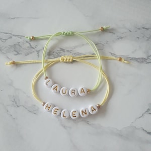 May include: Two friendship bracelets with the names "Laura" and "Helena" spelled out in gold letters on white beads. The bracelets are made with yellow and green string.