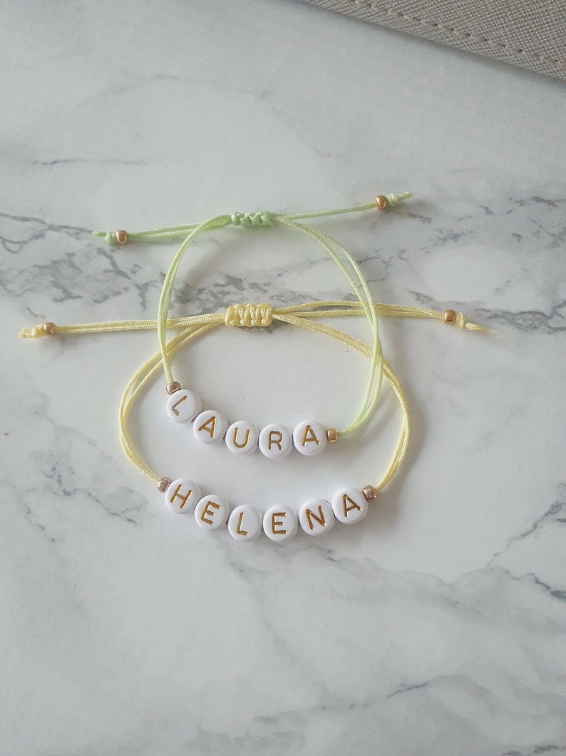 May include: Two friendship bracelets with the names "Laura" and "Helena" spelled out in gold letters on white beads. The bracelets are made with yellow and green string.