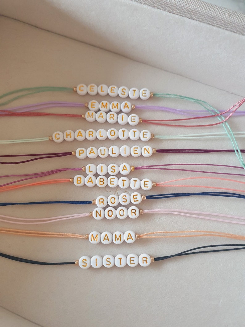 May include: A set of 10 colorful string bracelets with white beads spelling out different names, including Celeste, Emma, Marie, Charlotte, Paulien, Lisa, Babette, Rose, Noor, Mama, and Sister.