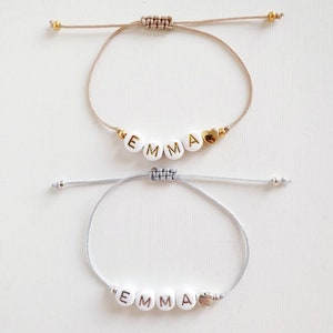 Può includere: Due braccialetti regolabili con il nome "EMMA" scritto in lettere dorate e argentate su perline bianche. Ogni braccialetto ha un piccolo ciondolo a forma di cuore dorato.