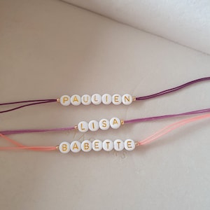 May include: Three bracelets with white beads and gold lettering spelling out the names "Paulien", "Lisa", and "Babette". The bracelets are on a white surface and are tied with different colored strings: purple, pink, and light pink.