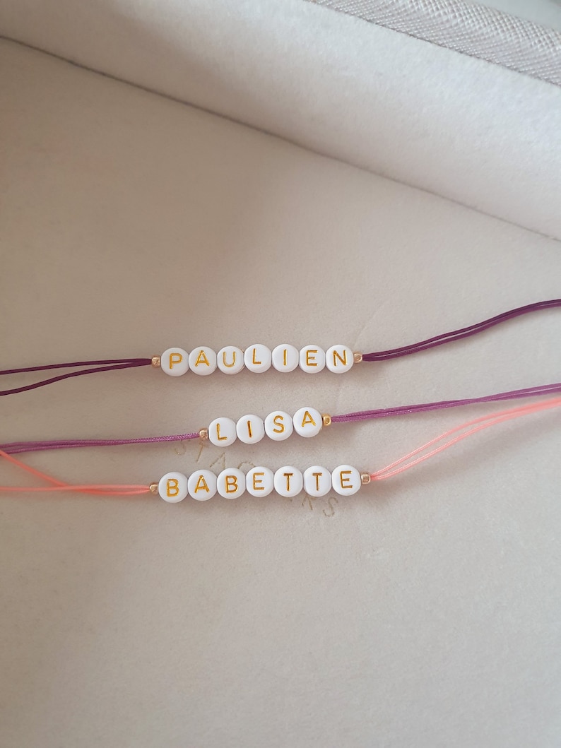 May include: Three bracelets with white beads and gold lettering spelling out the names "Paulien", "Lisa", and "Babette". The bracelets are on a white surface and are tied with different colored strings: purple, pink, and light pink.