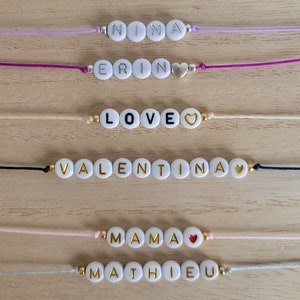 Op de afbeelding: Zes witte kralenarmbanden met gouden letters die de namen "NINA", "ERIN", "LOVE", "VALENTINA", "MAMA" en "MATHIEU" spellen. Elke armband heeft een klein gouden hartje.