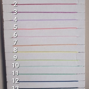 May include: A white board with 13 numbered lines. Each line has a different color string stretched across it. The colors are purple, pink, red, lavender, peach, light pink, beige, yellow, light green, dark green, blue, and black.