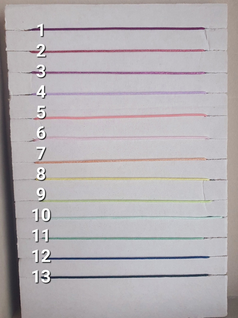 May include: A white board with 13 numbered lines. Each line has a different color string stretched across it. The colors are purple, pink, red, lavender, peach, light pink, beige, yellow, light green, dark green, blue, and black.
